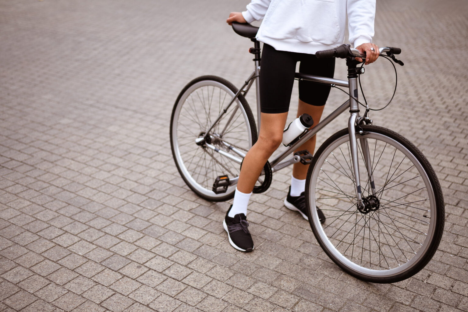 Faceless shot of african american woman in hoodie and cycling shorts standing near bicycle on street Faceless shot of african american woman in hoodie and cycling shorts standing near bicycle on street